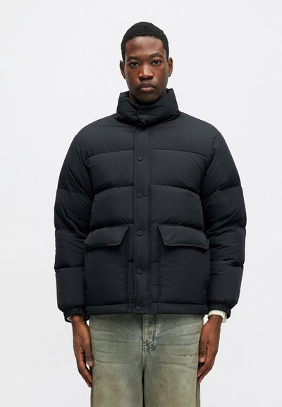 Vyras vilki juodą pūkinę striukę su snapeliais ir kišenėmis, derinamą su šviesiais, nudilusiomis džinsais, stovintis priešais vienodai baltą foną.