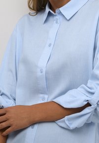 Lichtblauw knoopshirt met een kraag, gemaakt van een textuurstof. Heeft opgerolde mouwen en vijf knopen aan de voorkant.