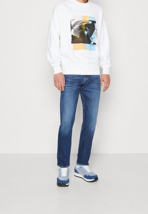 Felpa bianca con stampa grafica che presenta forme geometriche, abbinata a jeans blu sbiaditi e sneaker azzurre.