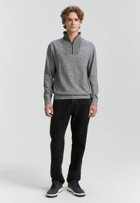 Grauer Pullover mit Reißverschluss und strukturierter Oberfläche, kombiniert mit schwarzen Hosen und Schuhen mit weißer Sohle. Modell steht aufrecht.