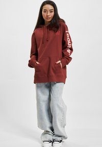 Felpa con cappuccio bordeaux, dotata di tasca e cordino, con testo del logo bianco sulla manica. Abbinata a jeans a gamba larga blu chiaro e sneakers nere.