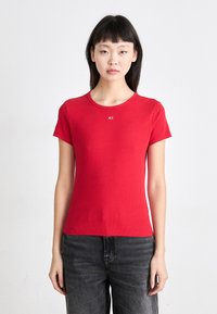 Camiseta de manga corta roja con un cuerpo ajustado, escote redondo y un pequeño detalle de logo en el pecho. Tela suave y elástica.