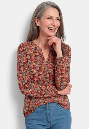 Femme d'âge moyen souriante aux cheveux gris, portant un haut à manches longues à motif floral et un jean bleu, debout, la main près du visage.