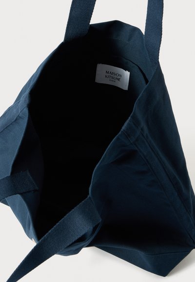 Σακούλα από καμβά σε ναυτικό μπλε χρώμα με ανθεκτικούς υφασμάτινους χειροκράτες και απλό σχέδιο. Στο εσωτερικό εμφανίζεται η ετικέτα: "MAISON KITSUNÉ PARIS."