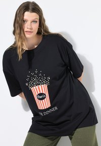 Mujer con pelo largo que lleva grandes pendientes de aro y una camiseta negra con un cubo de palomitas y el texto "Perlas para la Cena".