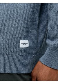 Grauer Strickpullover mit strukturiertem Muster, gerippten Bündchen und Saum, mit einem weißen Etikett, auf dem "JACK & JONES EST. 1990." steht.