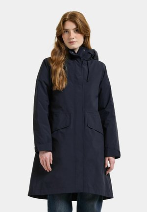 Woman with long red hair wearing a navy blue hooded parka coat and blue jeans standing against a plain light background.