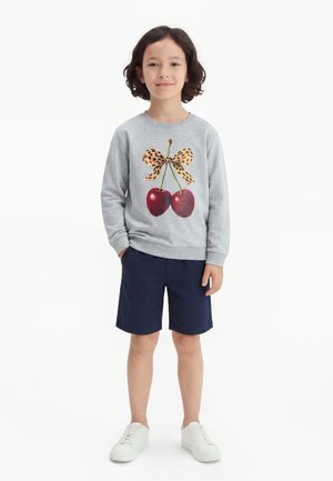 Enfant aux cheveux foncés portant un sweat-shirt gris avec un imprimé cerises et nœud léopard, un short bleu marine et des baskets blanches, debout les mains dans les poches.