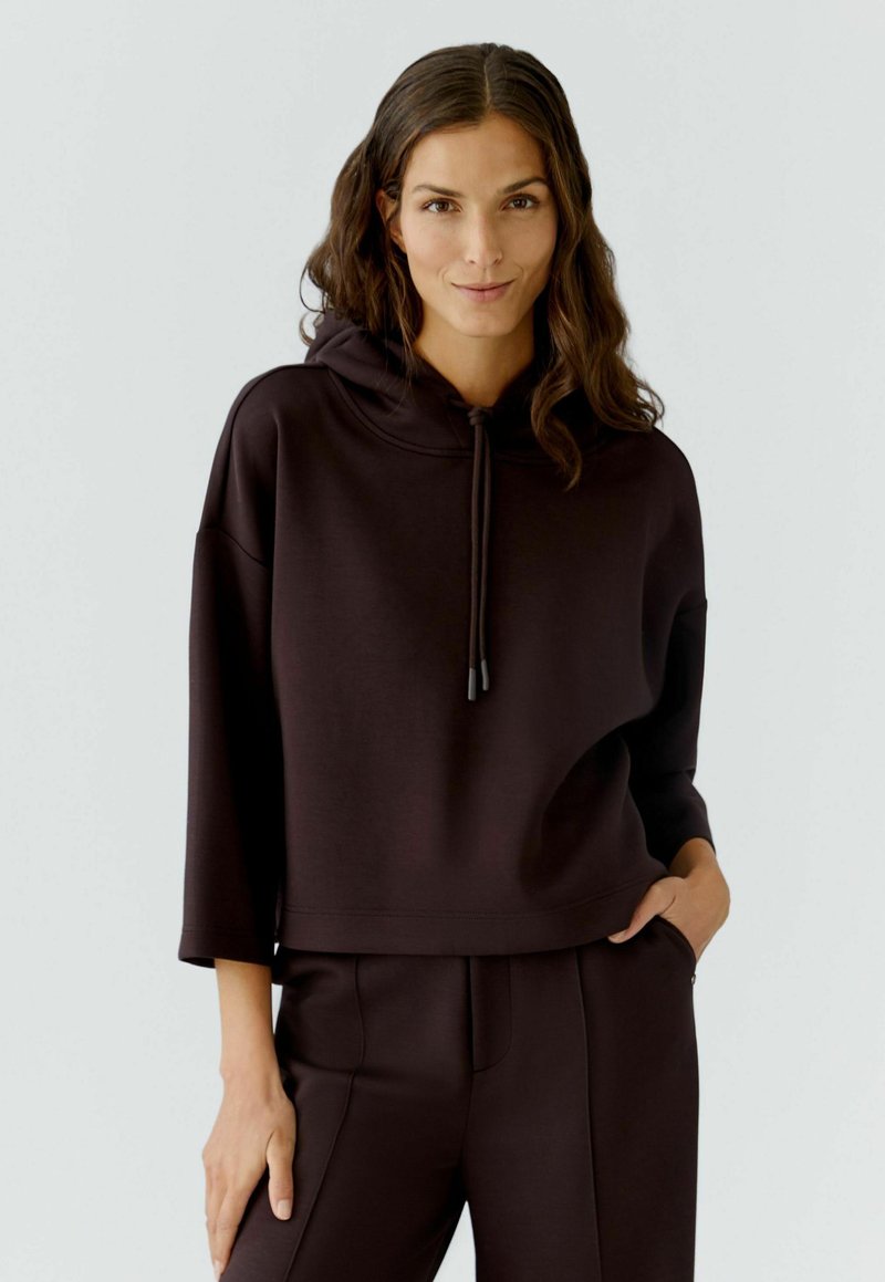 Braune, kurz geschnittene Kapuzenjacke mit Kapuze, Kordelzügen, weichem Material und weiten Ärmeln, kombiniert mit passenden Hosen für einen abgestimmten Look.