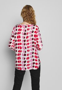 Blouse à manches longues avec une coupe ample, arborant un motif abstrait rose et rouge sur fond blanc. Tissu doux, encolure arrondie.