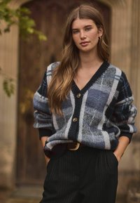 Cardigan à carreaux bleu et gris avec un col en V, de grands boutons et des poignets côtelés, porté sur un pantalon noir à rayures.