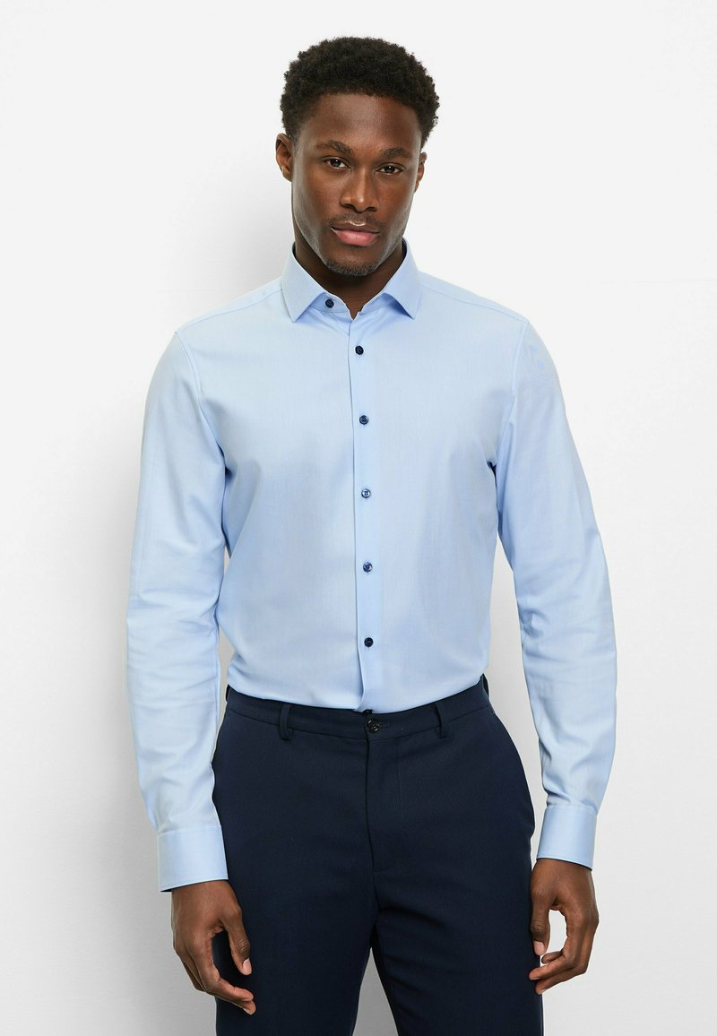 Hellblaues, langärmliges Hemd mit Knöpfen, figurbetontem Schnitt, dunklen Knöpfen und klassischem Kragen, getragen zu dunklen Hosen.