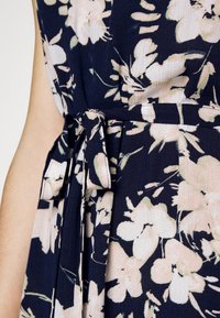 Szczegół sukienki w kwiaty z granatowym tłem i różowymi kwiatami; materiał jest lekki, z akcentem w postaci związanej talii i plisowaną teksturą.