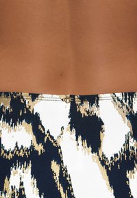 Top bez rukávov s abstraktným vzorom v námorníckej modrej, bielej a zlatej farbe s jemnou textúrou; obsahuje malú zlatú sponu na zadnej strane.