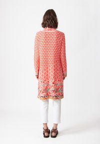 Garšīga, rakstaina tunika korallkrāsā ar ģeometriskiem dizainiem, piedurknēm līdz plaukstām un apakšmalu ar kontrastējošu maldēšanu. Valkāta virs baltām biksēm.