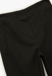 Pantalones ajustados negros con cintura alta, pliegues marcados y bolsillos laterales. La tela tiene una textura suave y un ajuste estructurado.