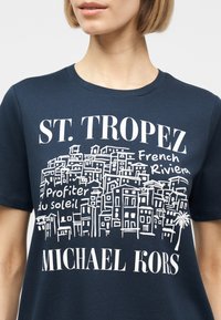 Γυναίκα που φοράει λευκό μπλουζάκι σε ναυτικό μπλε χρώμα με λευκό σκίτσο κτιρίων και κείμενο "ST. TROPEZ French Riviera Profiter du soleil" και "MICHAEL KORS."