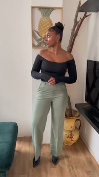 Sort off-shoulder bluse med lange ærmer, parret med lysegrønne højtaljede bukser og sorte spidse hæle. Trægulv og dekorativ vase.