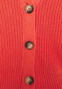 Korallfärgad stickad cardigan med ett texturerat mönster, en knappslå med fyra stora mörka knappar och ribbade muddar vid ärmarna och fållen.