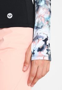 Top sportivo nero con unPattern floreale colorato sulle maniche lunghe. Tessuto morbido con un design aderente. Mano appoggiata su un pantalone rosa.