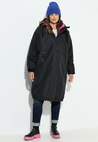 Parka negra con forro rosa, bolsillos con cremallera y cordón ajustable en la cintura y la capucha. Combinada con jeans oscuros y botas con suela rosa.