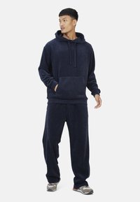 Mężczyzna w granatowym, rypsowym hoodie i dopasowanych szerokich spodniach, w szarych i beżowych sneakersach, stoi na tle białym.