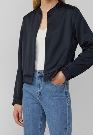 Kvinde iført en sort satinjakke uden krave over en hvid top og lyseblå jeans med hånden i lommen foran en ensfarvet baggrund.