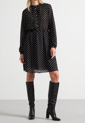 Kobieta w czarnej sukience do kolan w białe groszki z długimi rękawami, w połączeniu z czarnymi, sięgającymi kolan butami na obcasie.