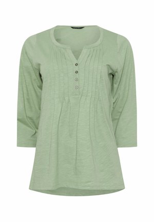 Camicia lunga a maniche lunghe verde chiaro con scollo rotondo, pattina con quattro bottoni e pieghe verticali lungo il davanti.