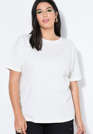 Kvinde med langt mørkt hår iført hvid t-shirt med små perleaccenter, sorte bukser, gyldne creoløreringe og grøn øjenskygge.