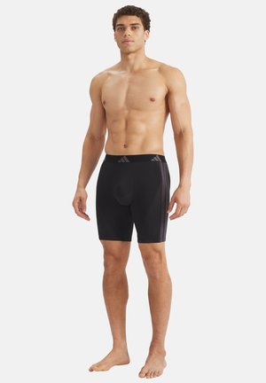 Athletischer junger Mann, barfuß stehend, ohne Hemd, trägt schwarze Kompressionsshorts bis zum Knie mit seitlichen Streifen und einem markierten Taillenbund.