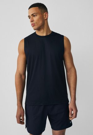 Muskelöser Mann steht, trägt ein ärmelloses schwarzes Sportshirt und schwarze Shorts, schaut nach links vor einem schlichten Hintergrund.