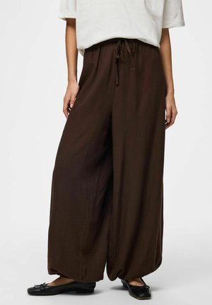 Kobieta nosząca luźne, ciemnobrązowe szerokie spodnie ze sznurkiem w talii, w połączeniu z białym topem i czarnymi balerinami.