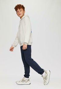 Lichtgrijze hoodie, marineblauwe trainingbroek en witte sneakers met grijze accenten. Model in een looppose tegen een effen achtergrond.