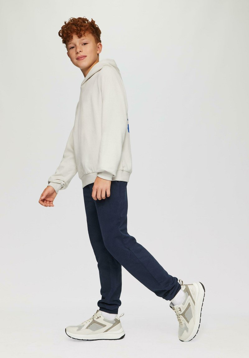 Lichtgrijze hoodie, marineblauwe trainingbroek en witte sneakers met grijze accenten. Model in een looppose tegen een effen achtergrond.