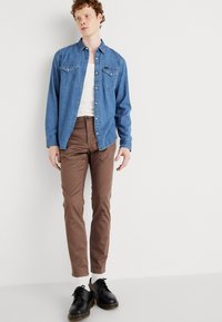 Denim button-up shirt in light blue, brown pants, black shoes with yellow accents, worn over white undershirt, standing against a neutral backdrop.