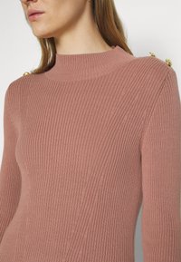 Close-up of a woman wearing a dusty pink ribbed sweater with gold buttons on the shoulder and long sleeves.