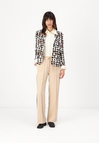 Zwarte blazer met een hartpatroon in beige, wit en rood; gecombineerd met beige broek met trekkoord en zwarte zijkanten; witte shirt eronder.