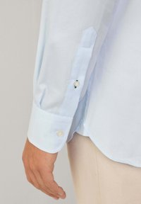 Camicia color blu chiaro con una texture a righe sottili, caratterizzata da due bottoni ai polsini e un orlo arrotondato. Il tessuto appare liscio e morbido.