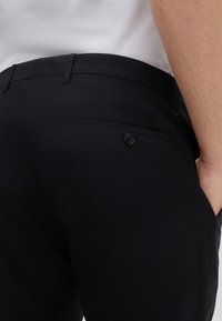Zwarte broek met een gladde textuur, slim fit en een achterzak met een zwarte knoop. Tailleband detail en subtiele stiksels zichtbaar.