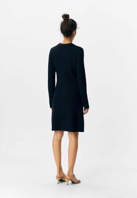 Rippstrickkleid in dunkelblau mit langen Ärmeln, Rundhalsausschnitt und gerader Silhouette, endend auf Oberschenkelhöhe. Kombiniert mit beigen, hohen Sandalen.