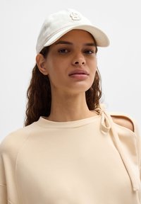 Cappello da baseball bianco con logo ricamato, indossato con una maglia beige chiara con scollatura ampia e un dettaglio con fiocco sulla spalla.