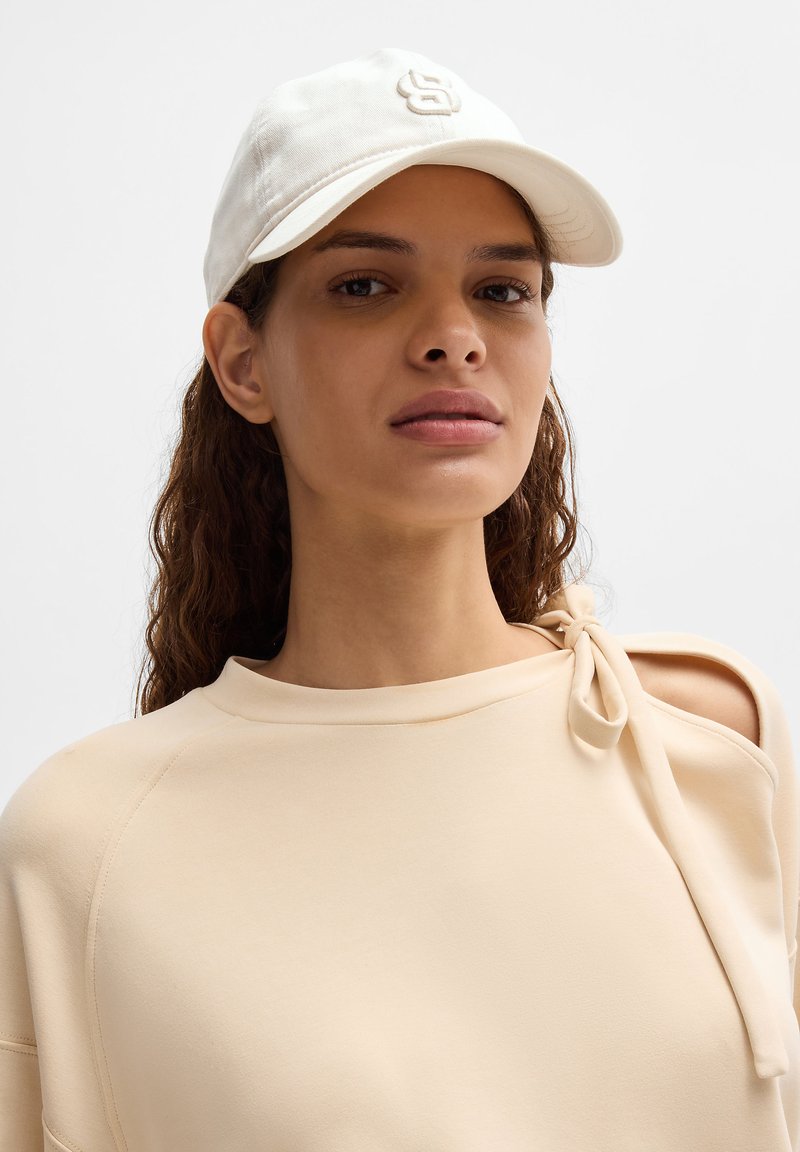 Casquette de baseball blanche avec logo brodé, portée avec un haut beige clair à encolure large et un accent de lien à l'épaule.