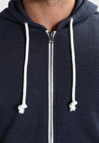 Sudadera con cremallera en color azul marino con cordones blancos y cremallera de metal. Fabricada en un tejido suave y texturizado. Cuenta con una capucha estándar y apertura frontal.