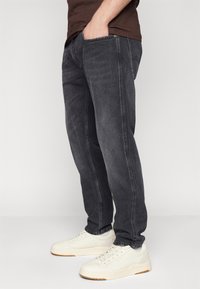 Jean en denim gris foncé avec des coutures visibles, poches avant et coupe droite, associés à des baskets blanches avec des détails perforés.