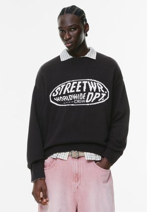 Dark knit sweater with a white graphic logo, layered over a light plaid shirt, paired with light pink denim pants.
