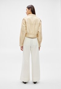 Sieviete ar gariem tumšiem matiem, ģērbusies bēša krāsas īsā jaciņā un augsta vidukļa plaša stila baltās biksēs, redzama no aizmugures.