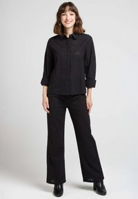 Chemise noire à manches retroussées, assortie à un pantalon de la même couleur. Les deux pièces sont en tissu texturé et à motifs, complétées par des bottines noires à la cheville.