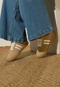 Persona che indossa sneakers beige con strisce bianche e pantaloni di jeans blu larghi, in piedi su un tappeto intrecciato contro un muro bianco.
