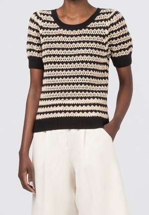 Vrouw die een zwart-beige gestreept, kortärmelig gebreid shirt draagt en een hooggesneden, wijde broek in crèmekleur met één hand in de zak.